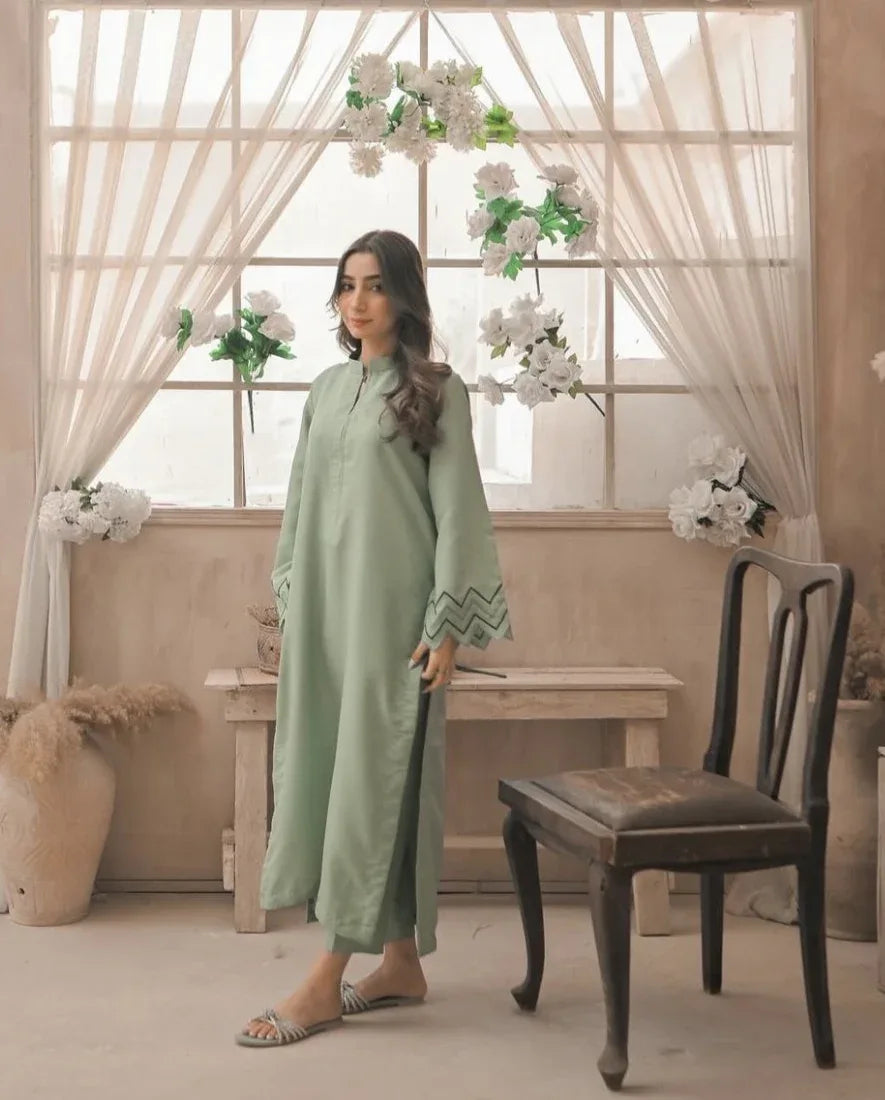 A Woman wearing an apple green dress with embroidery on sleeves and zigzag cuffs, standing beside a chair and against a window, smiling confidently. 