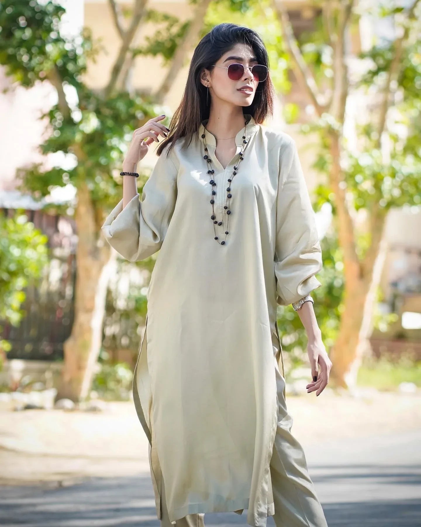 A women wearing a beige dress kurta  standing outdoors with a confident pose. Background is leafy and sunlit.