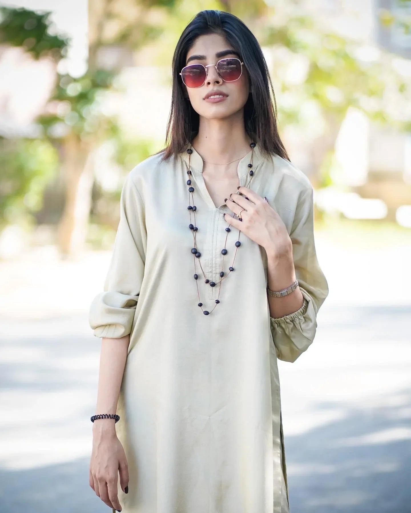 A women wearing a beige dress kurta  standing outdoors with a confident pose. Background is leafy and sunlit.