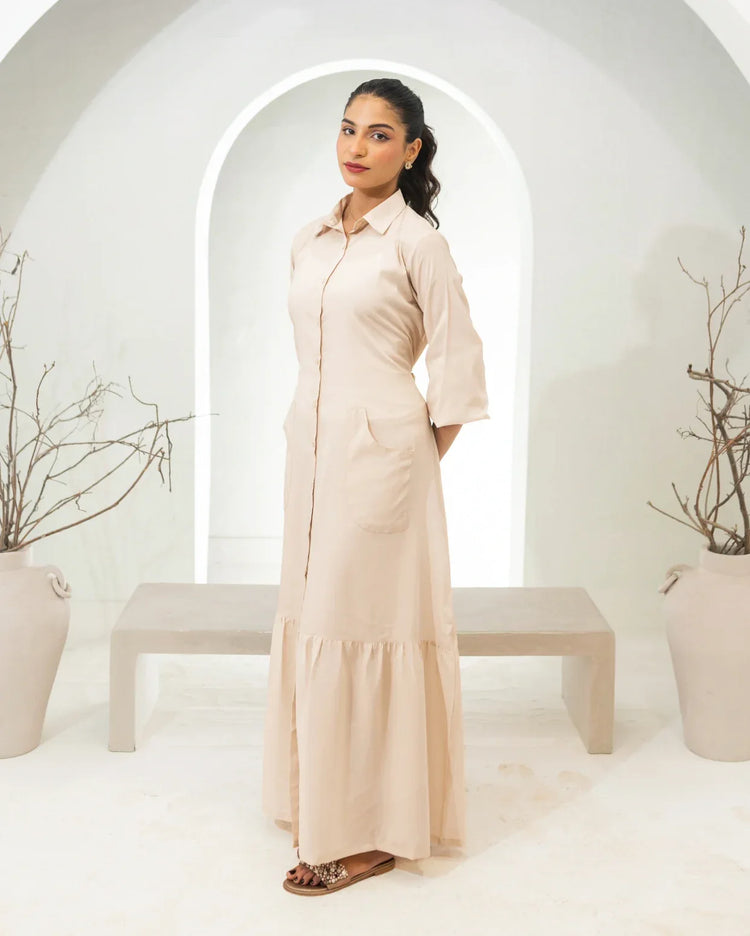 Woman wearing a long fawn dress standing in a minimalistic setting with white arches and potted plants.