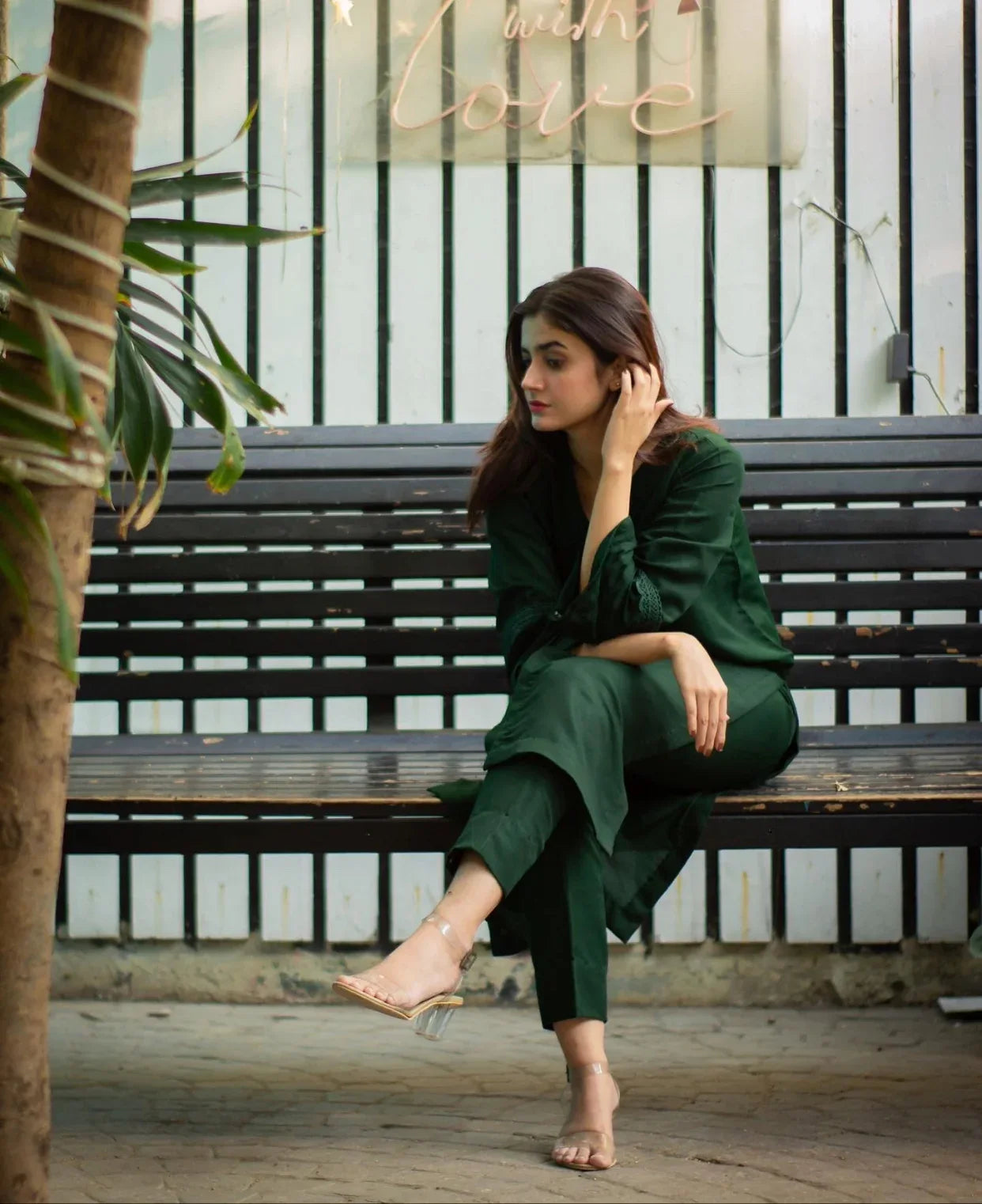 Woman in a bottle green outfit sitting on a bench with plants and a decorative sign in the background