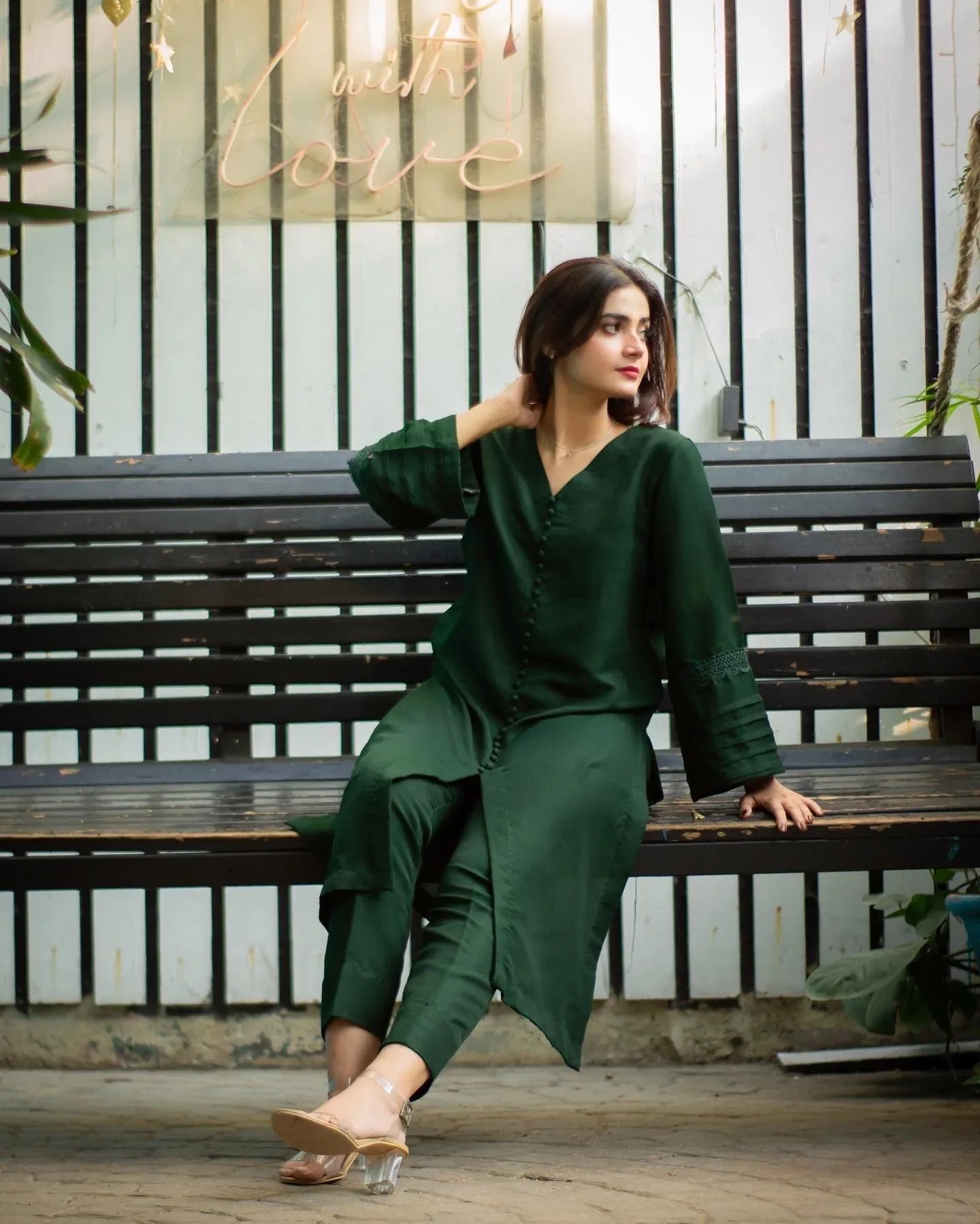 Woman in a bottle green outfit sitting on a bench with plants and a decorative sign in the background.