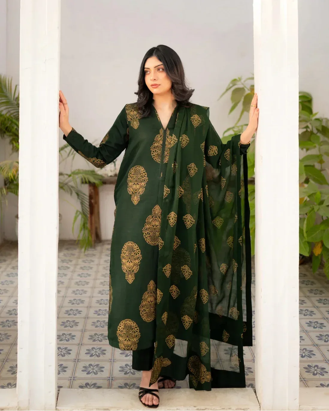 Woman wearing a bottle green traditional three piece dress with gold patterns in a room with plants.