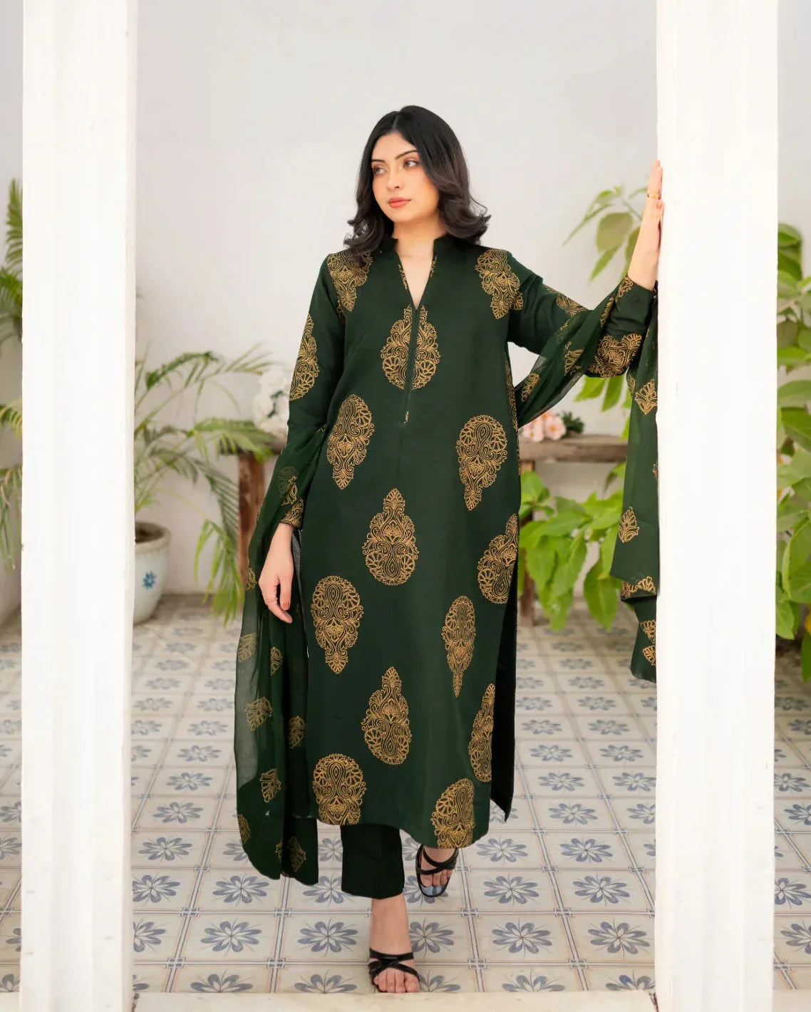 Woman wearing a bottle green dress with gold patterns in a room with plants and patterned tiles.