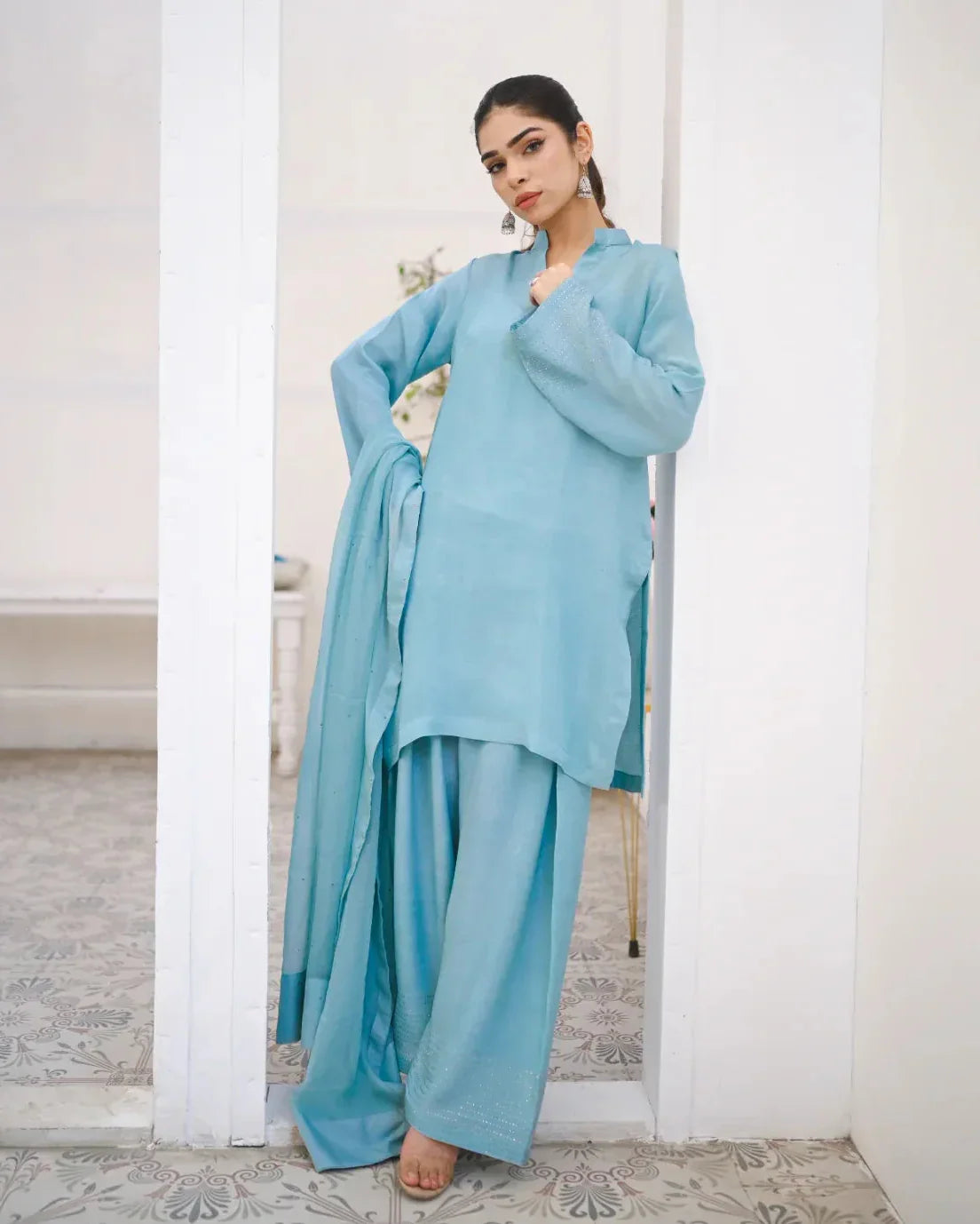 A woman wearing a Sky Blue three piece rawsilk dress which includes a farshi shalwar kameez with chiffon dupatta. The dress is embellished with stone details.