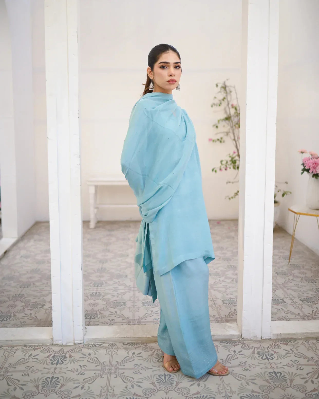 A woman wearing a Sky Blue three piece rawsilk dress which includes a farshi shalwar kameez with chiffon dupatta. The dress is embellished with stone details.