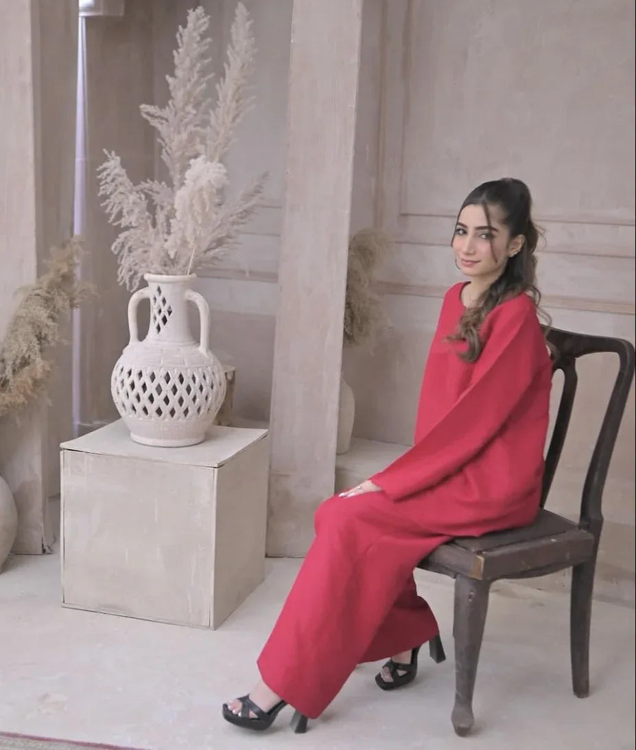 A woman in a deep pink outfit sitting on a chair against a beige, textured wall. She has a calm expression and wears black heels.