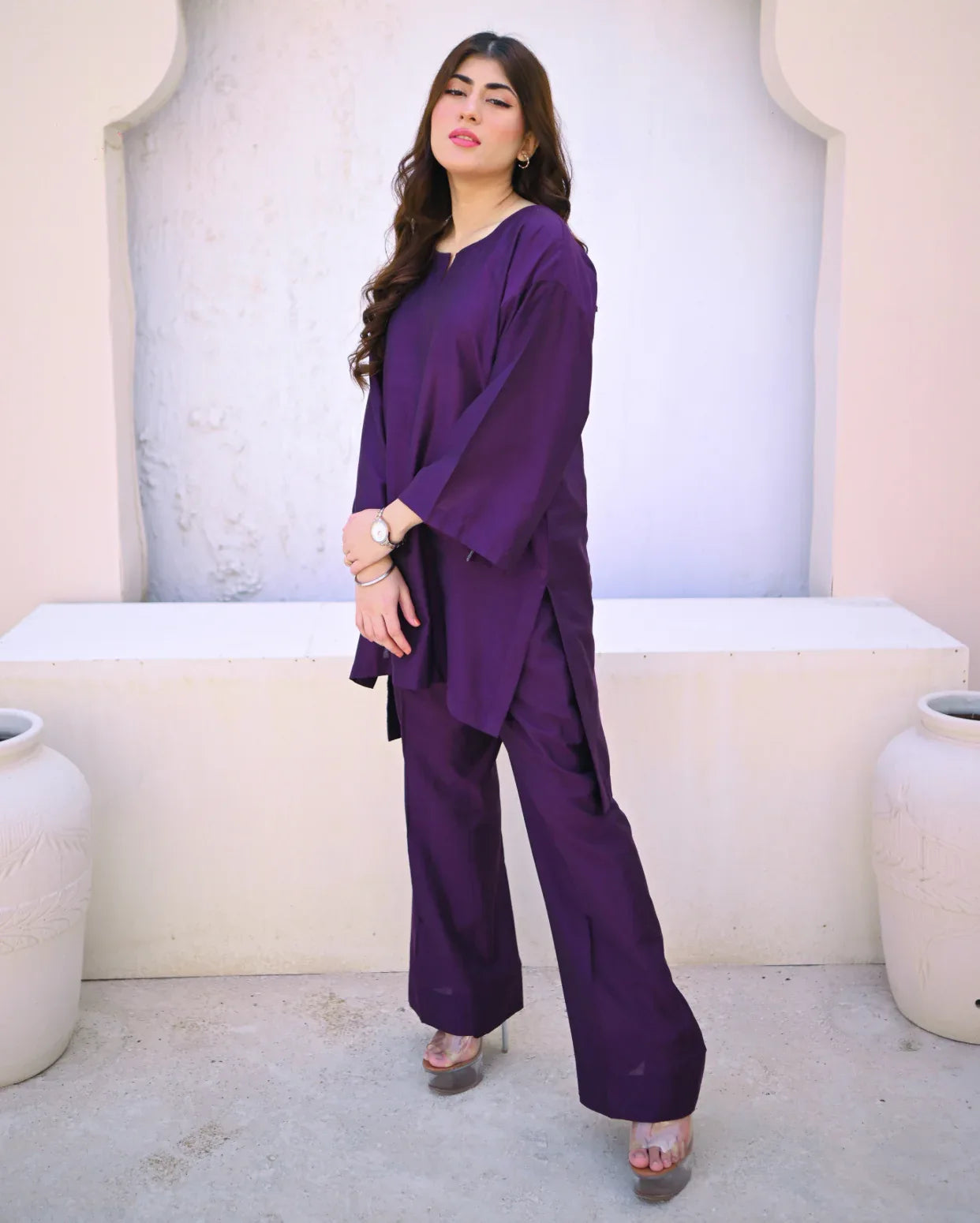 Woman wearing a purple outfit standing against a light-colored wall with decorative elements.