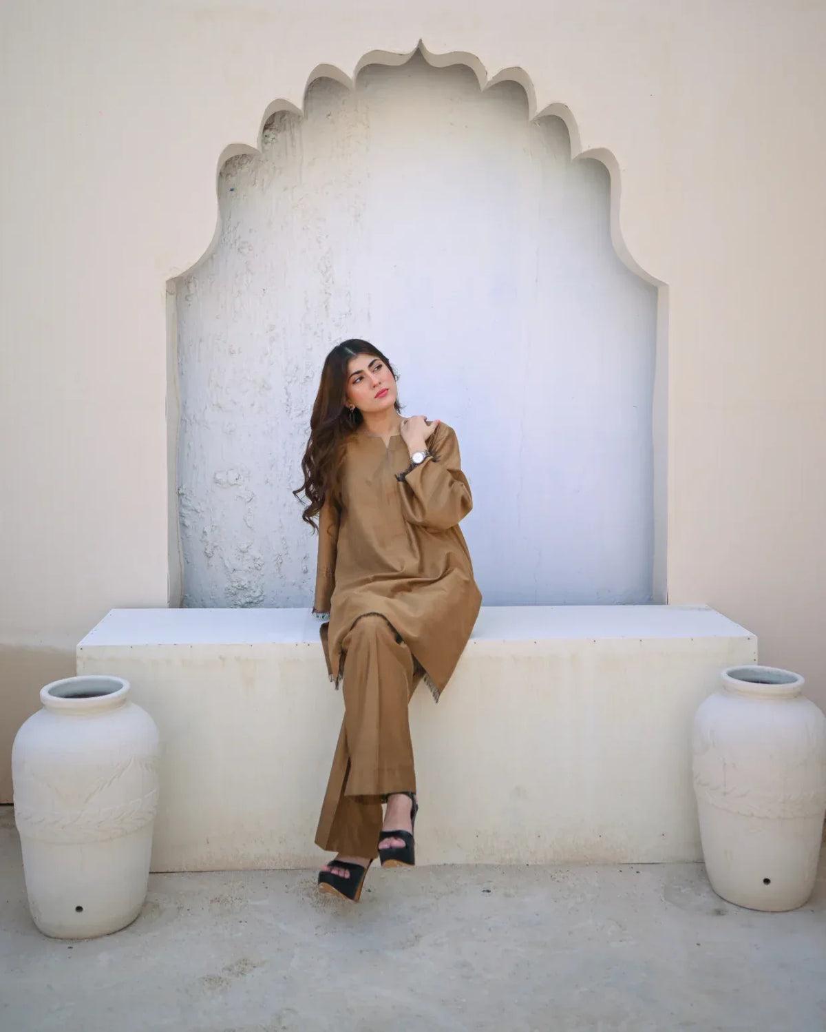 Woman in a beige outfit sitting on a white bench with decorative arch and vases.