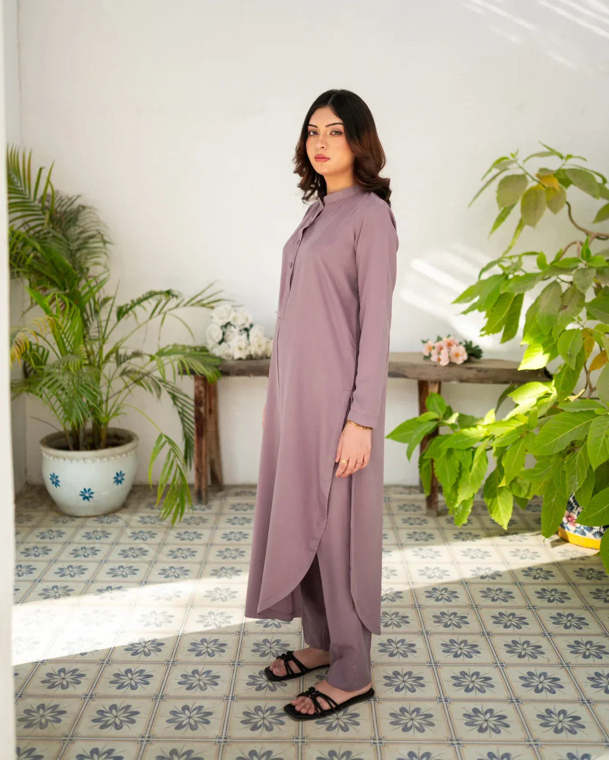 Woman in a long and lavender color dress standing in a room with plants and a tiled floor.