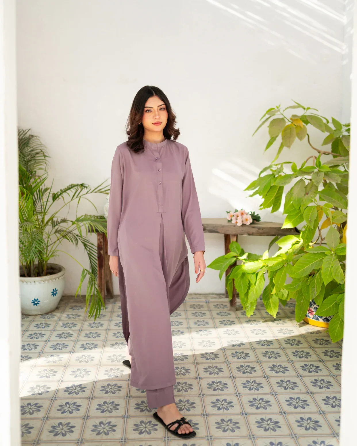 Woman in a lavender outfit standing in a room with plants and a tiled floor.