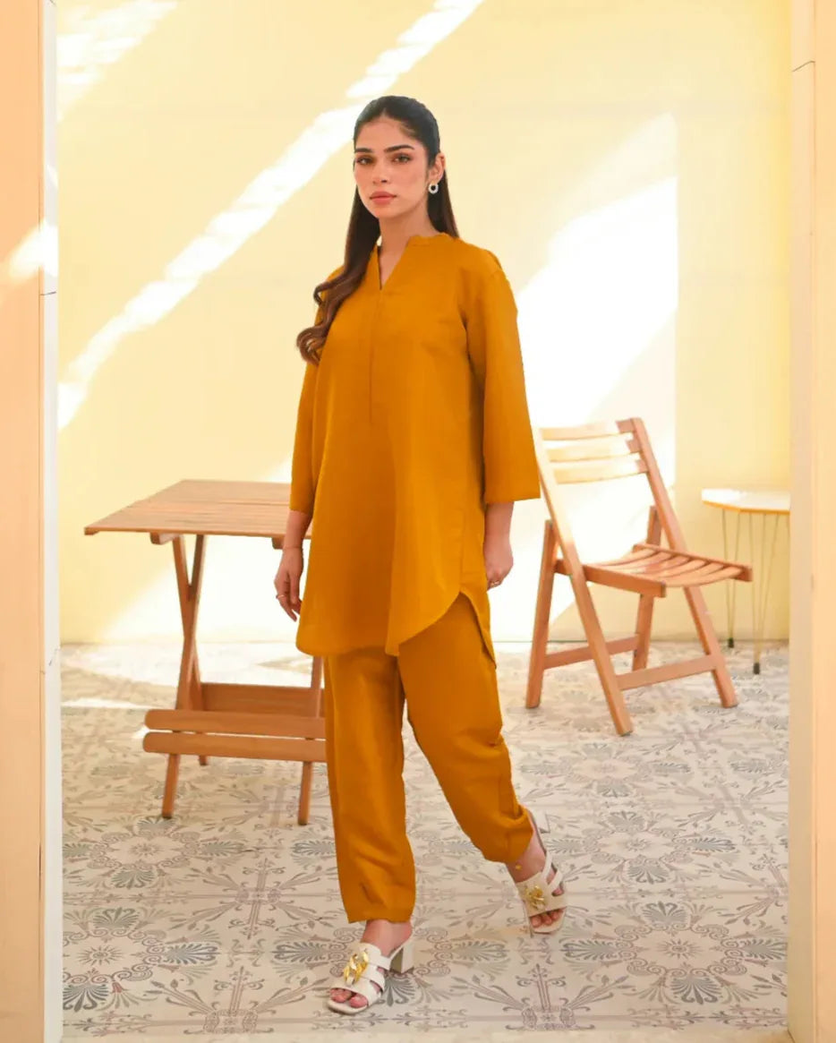 A woman wearing a mustard yellow two-piece dress with long sleeves, consisting of a shirt and trousers, standing in a sunny area next to a wooden bench.