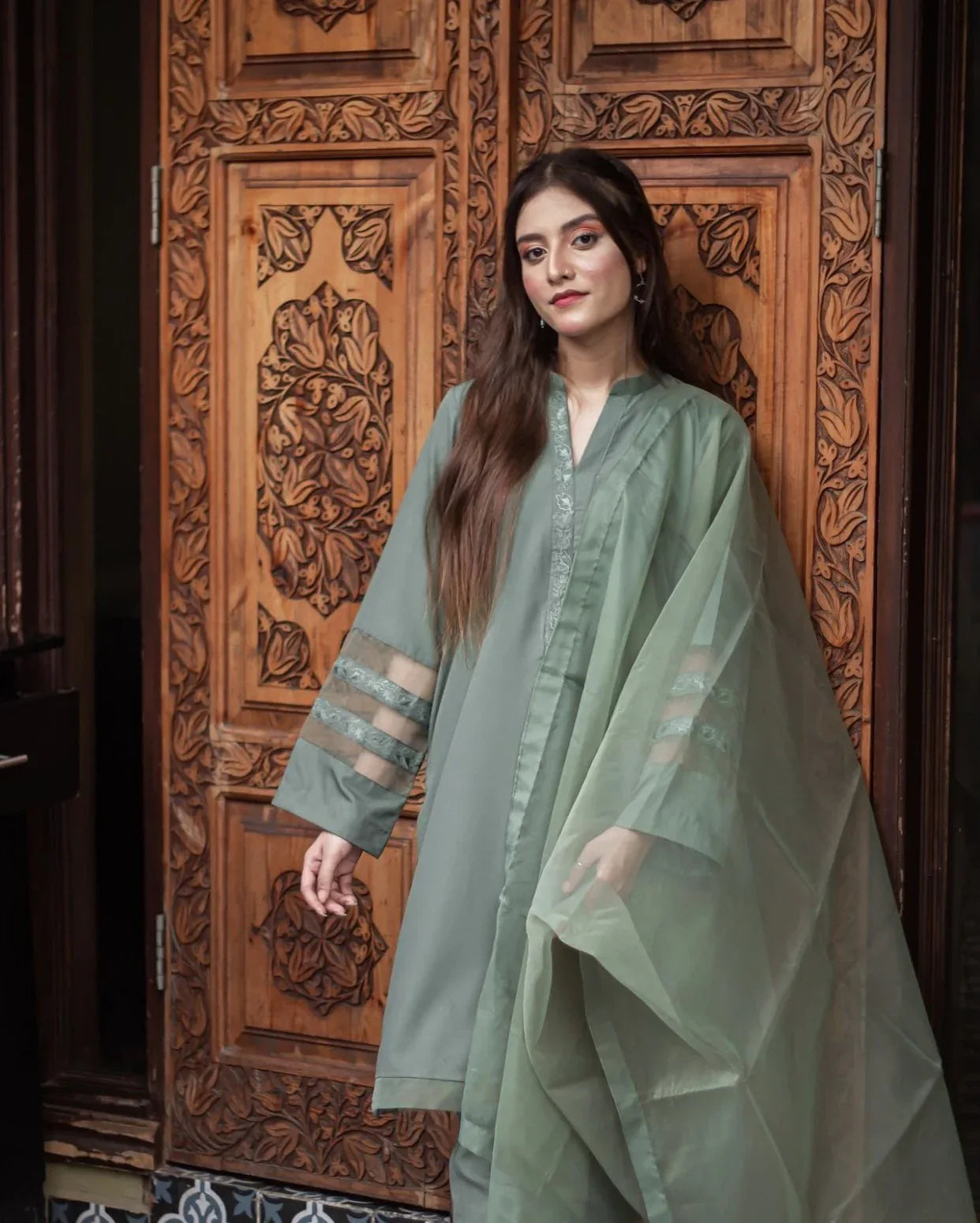 A woman in an olive green stitched dress stands confidently against an ornately carved wooden door, conveying elegance and cultural richness.
