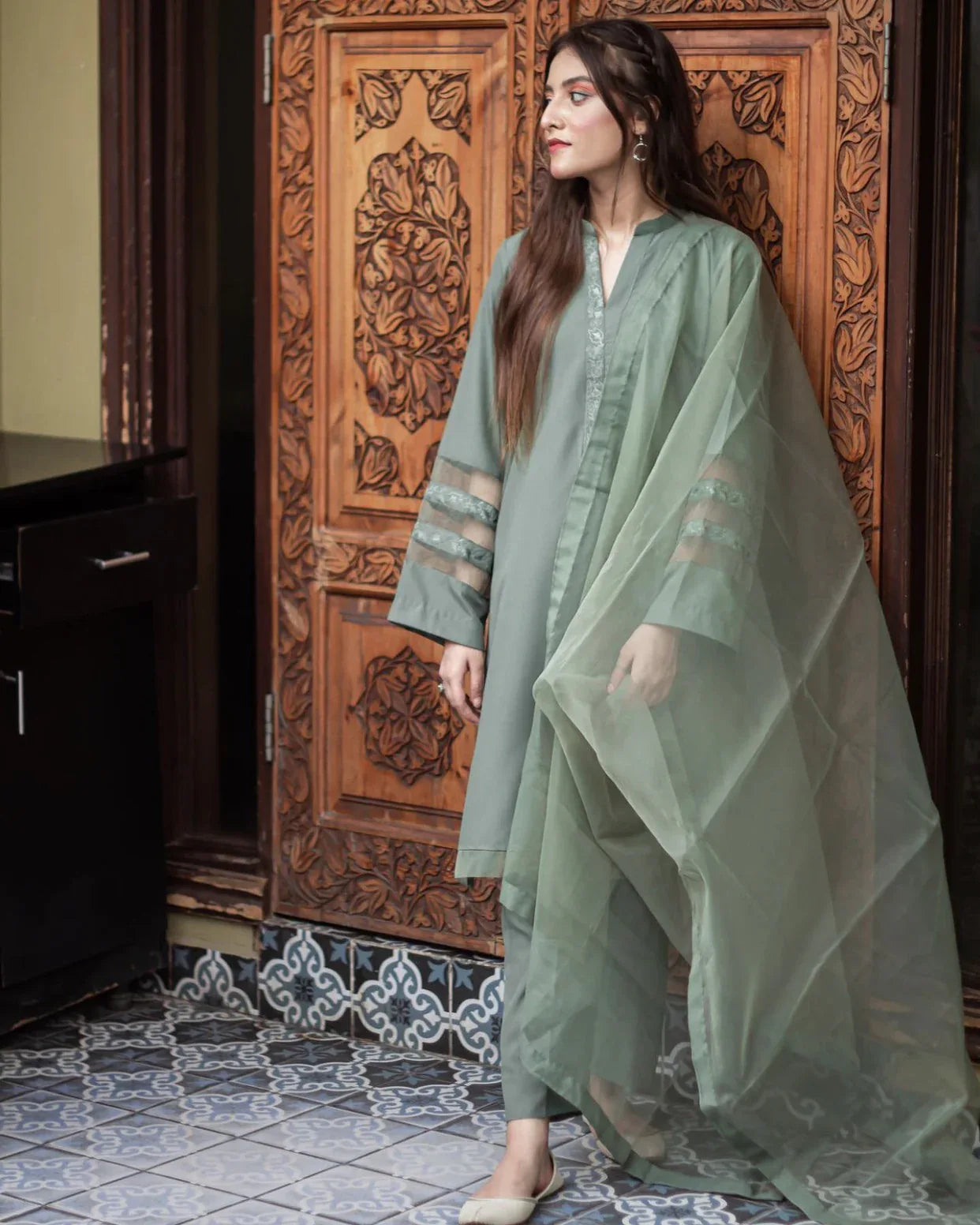 A woman in an olive green stitched dress stands confidently against an ornately carved wooden door, conveying elegance and cultural richness.