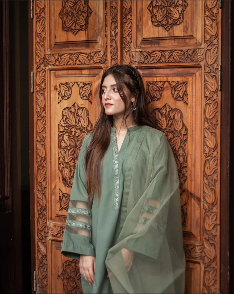 A woman in an olive green stitched dress stands confidently against an ornately carved wooden door, conveying elegance and cultural richness.