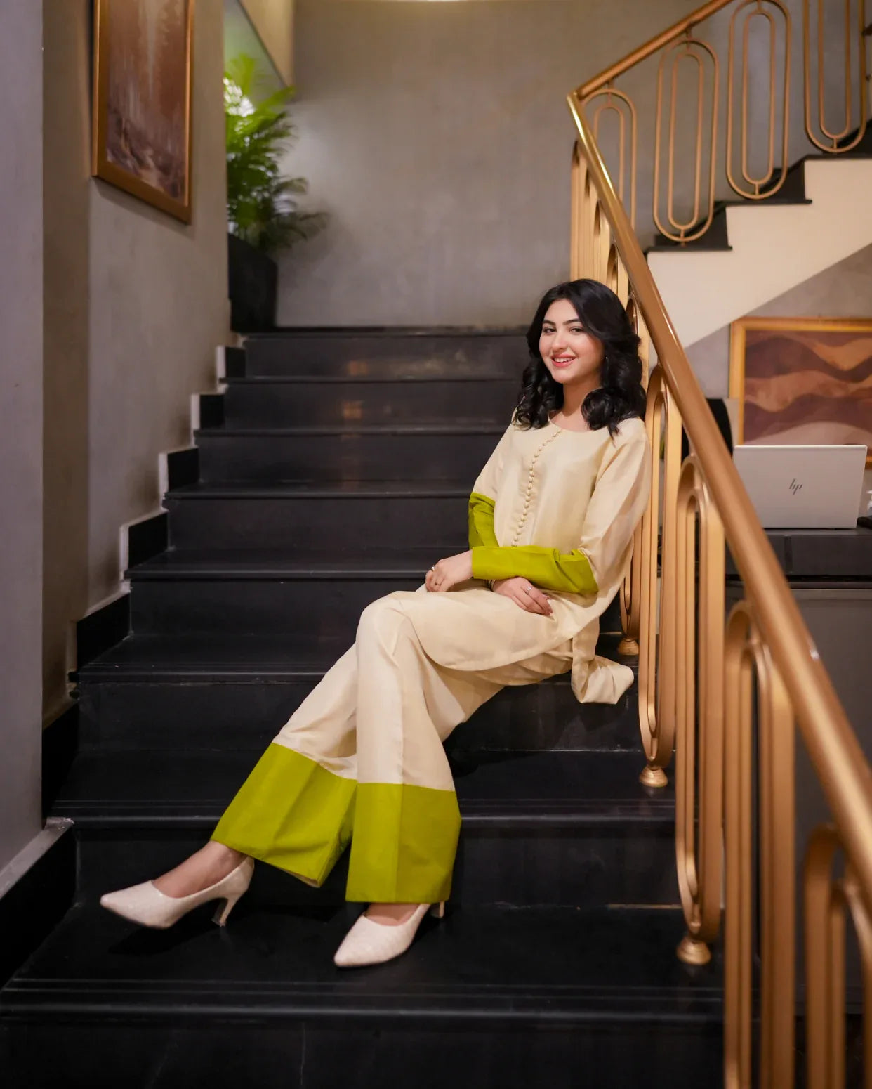 Woman in a cream and green outfit sitting on a staircase with gold railings.