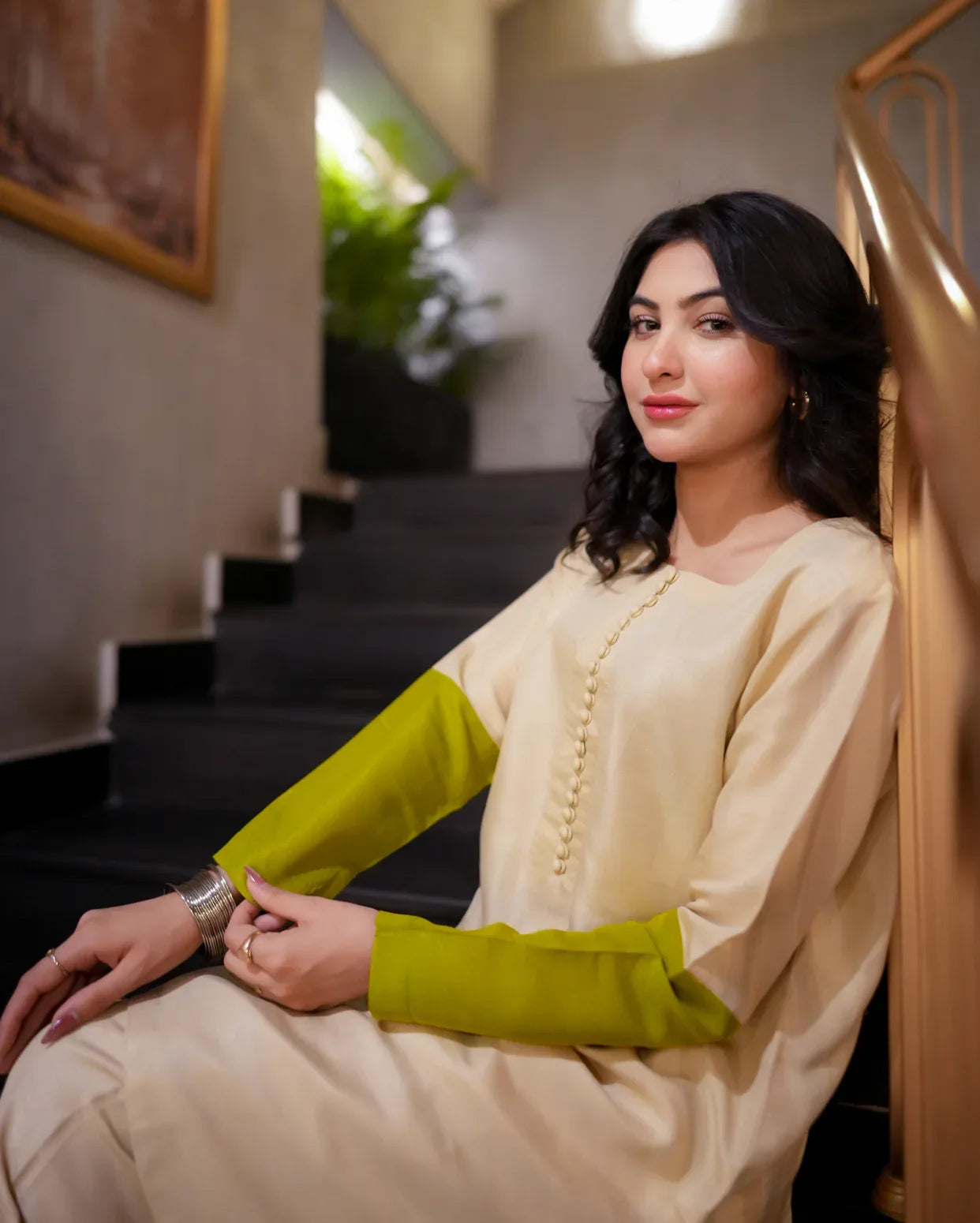 Woman in a creme color traditional outfit with half green sleeves sitting on a staircase.