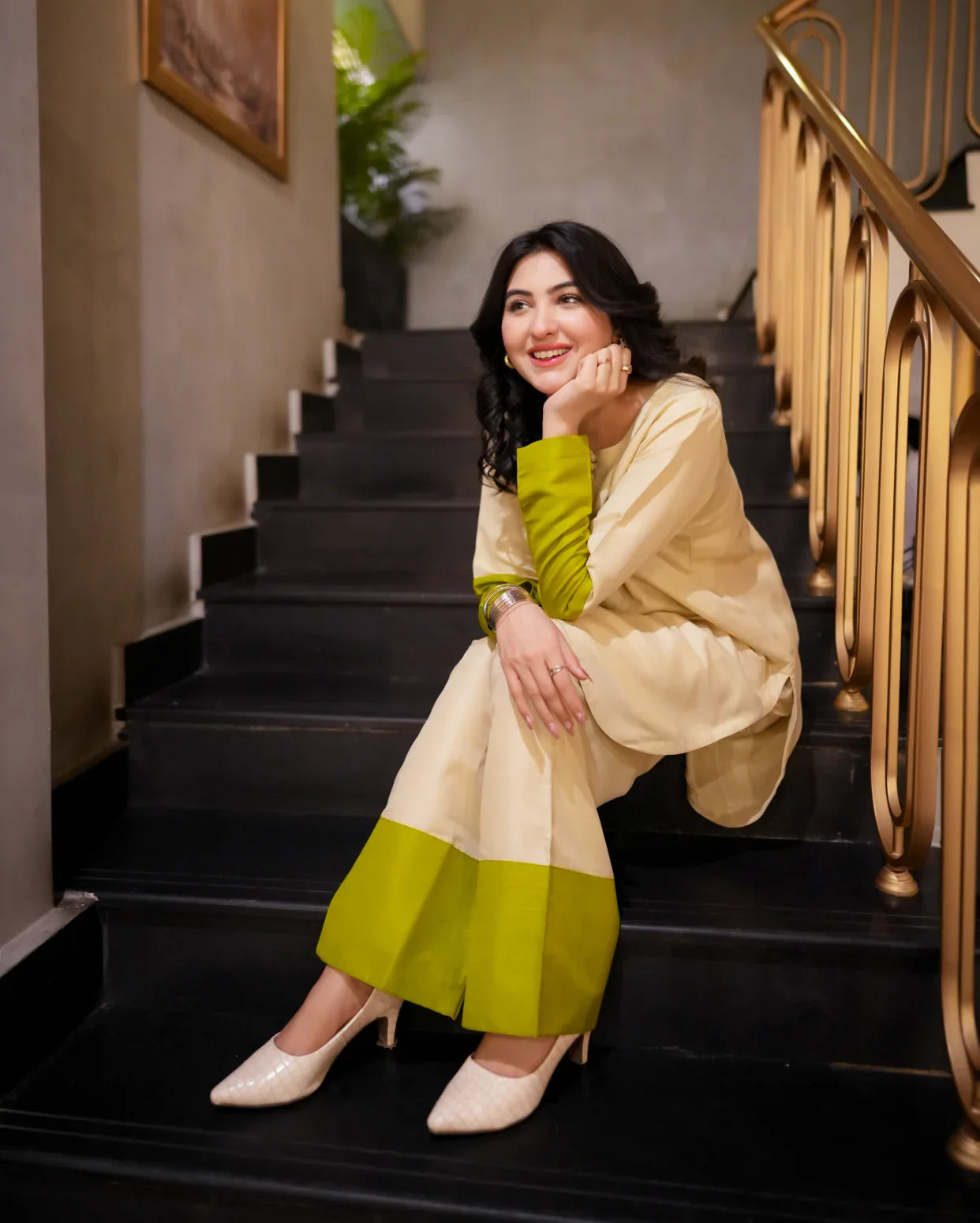 Woman sitting on a staircase wearing a green and beige outfit with a gold railing.