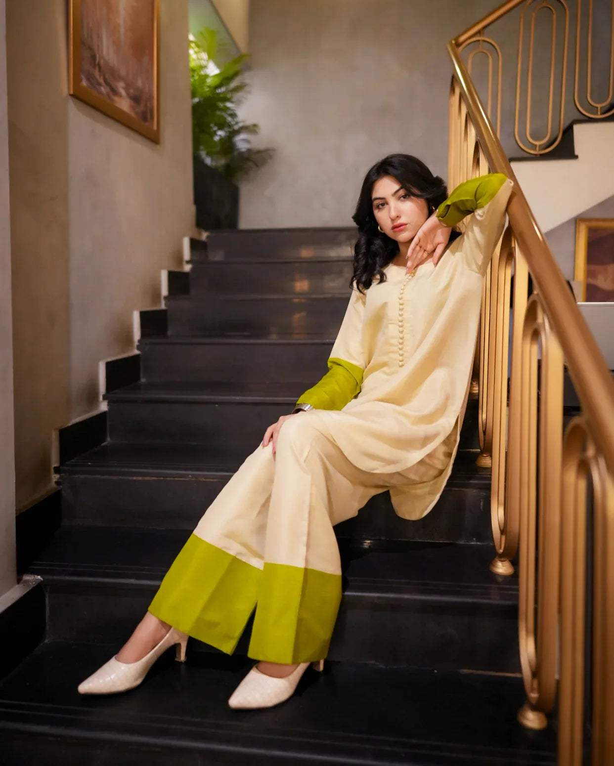 Woman in a cream and green outfit sitting on a staircase with gold railings.