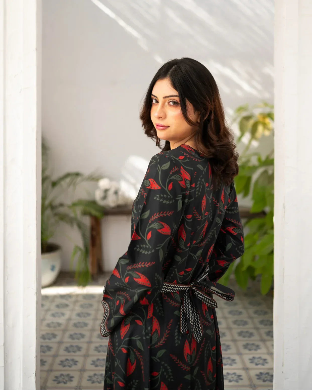 Woman wearing a black printed dress in a room with plants and a mirror.