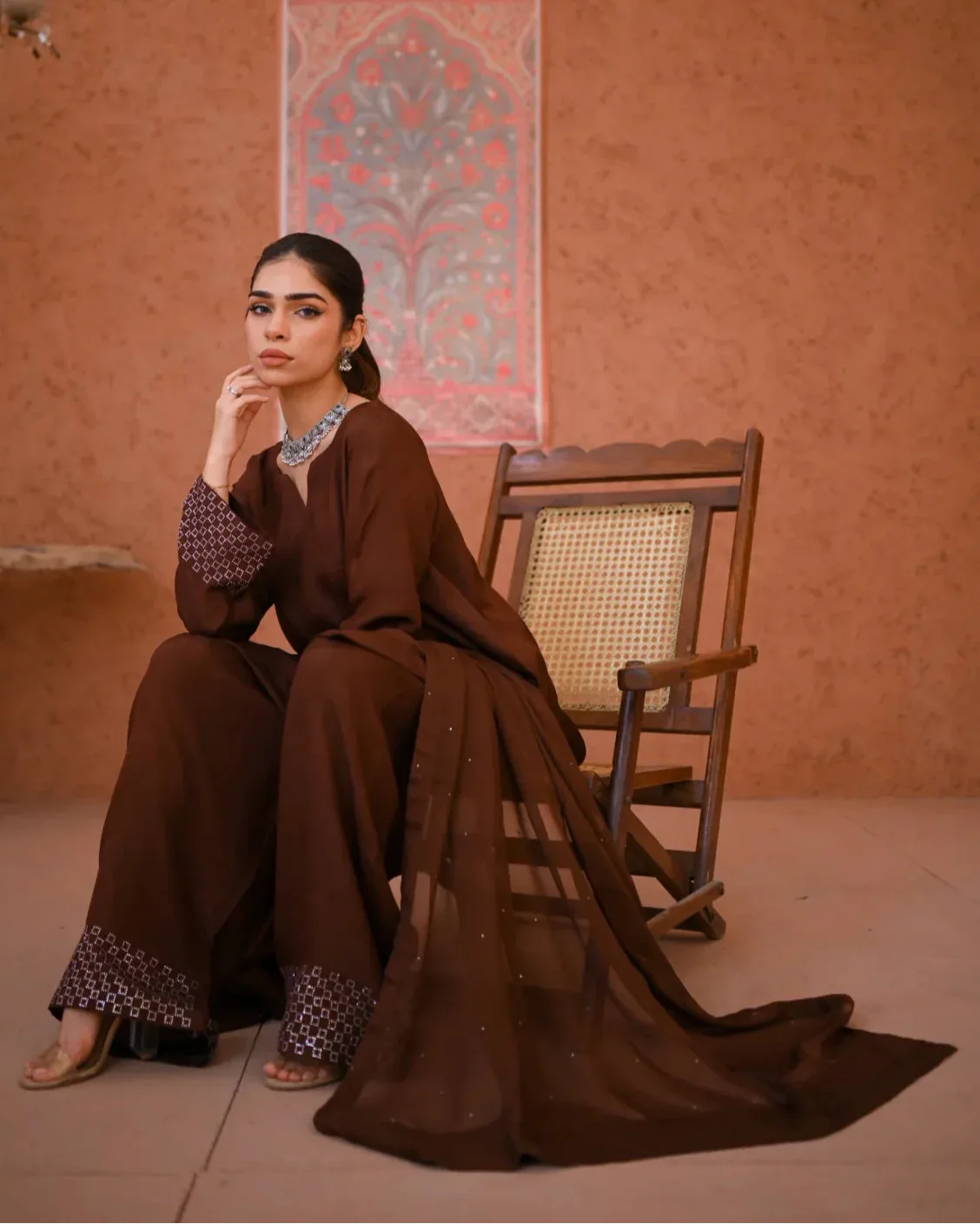 A woman wearing a brown three-piece rawsilk dress which includes a farshi shalwar kameez along with a chiffon dupatta. The dress is embellished with stone details.