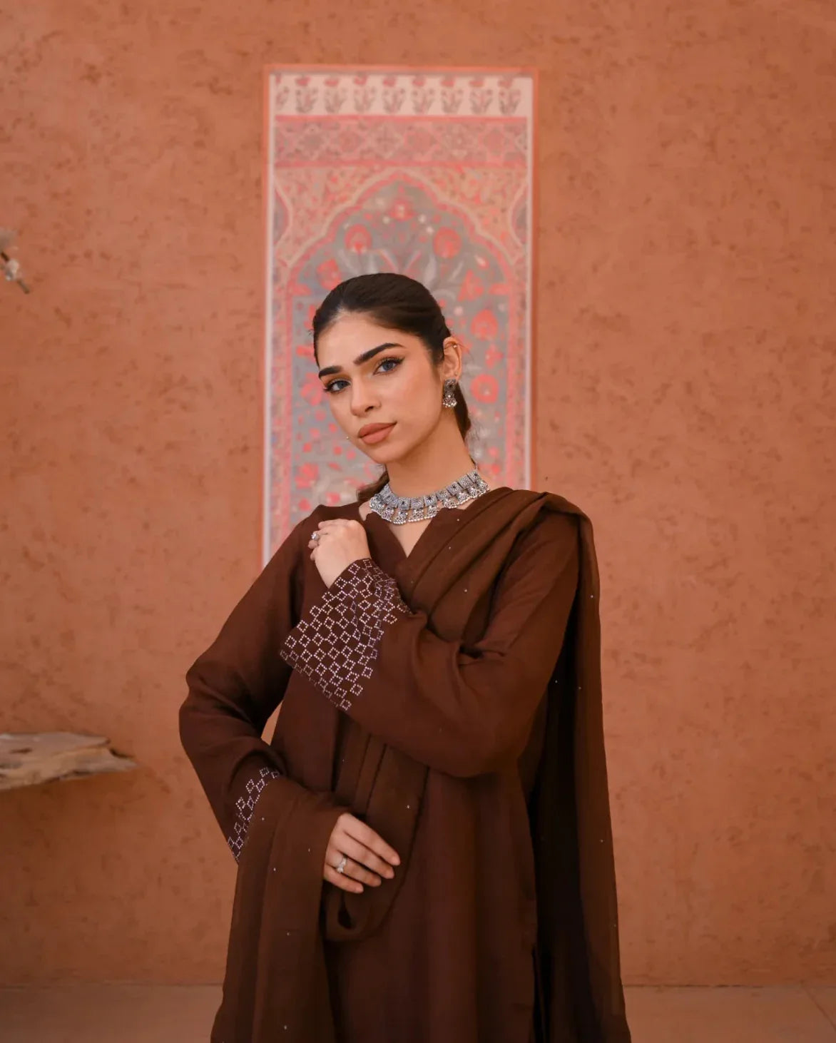 A woman wearing a brown rawsilk dress which includes a shirt and  a chiffon dupatta. The dress is embellished with stone details.