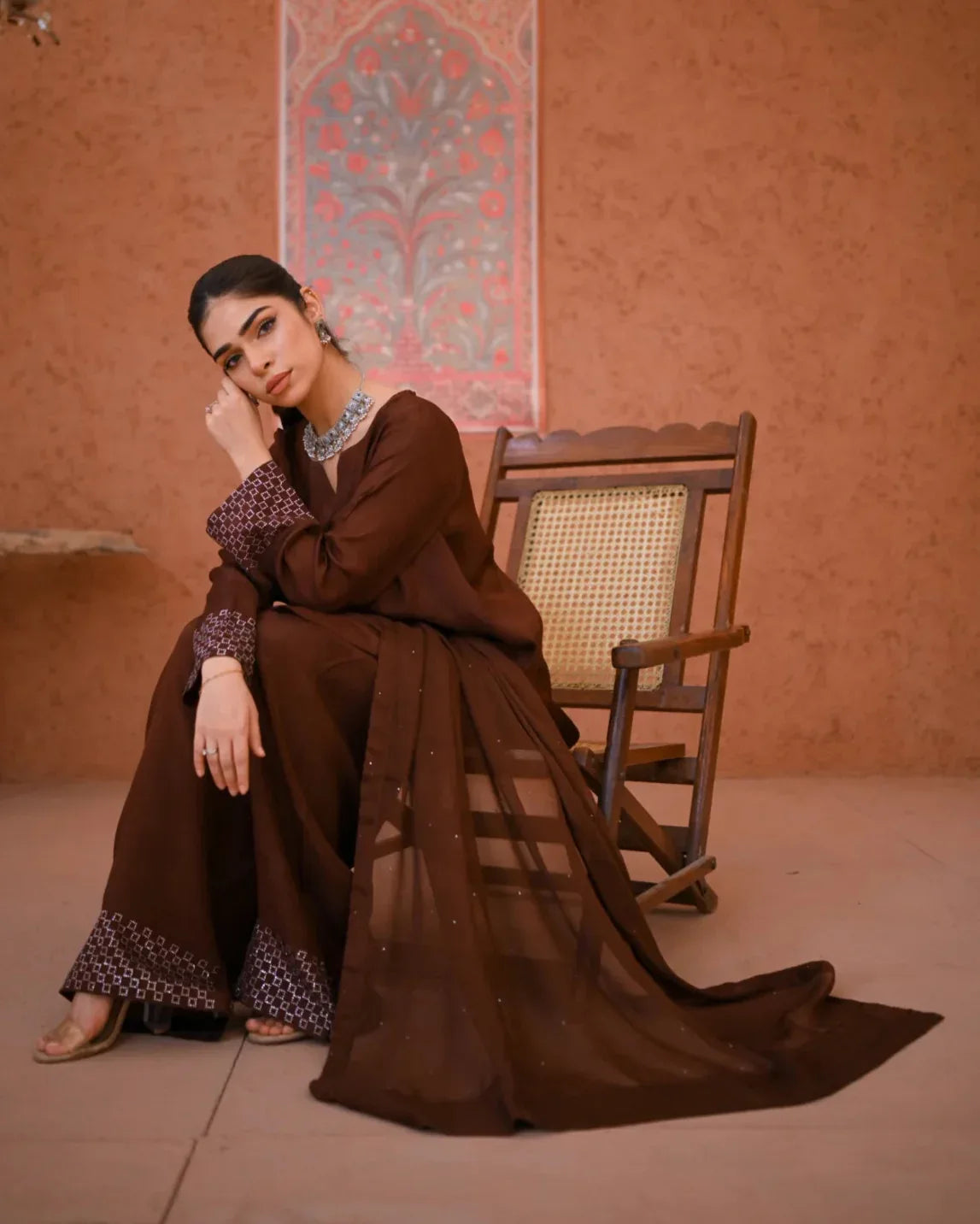 A woman wearing a brown three-piece rawsilk dress which includes a  farshi shalwar kameez along with a chiffon dupatta. The dress is embellished with stone details.