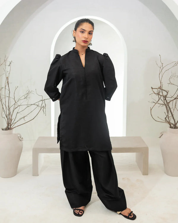 Woman wearing a black traditional two piece farshi shalwar kameez in a minimalistic setting with white arches and potted plants.