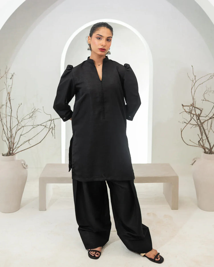 Woman wearing a black traditional two piece farshi shalwar kameez in a minimalistic setting with white arches and potted plants.