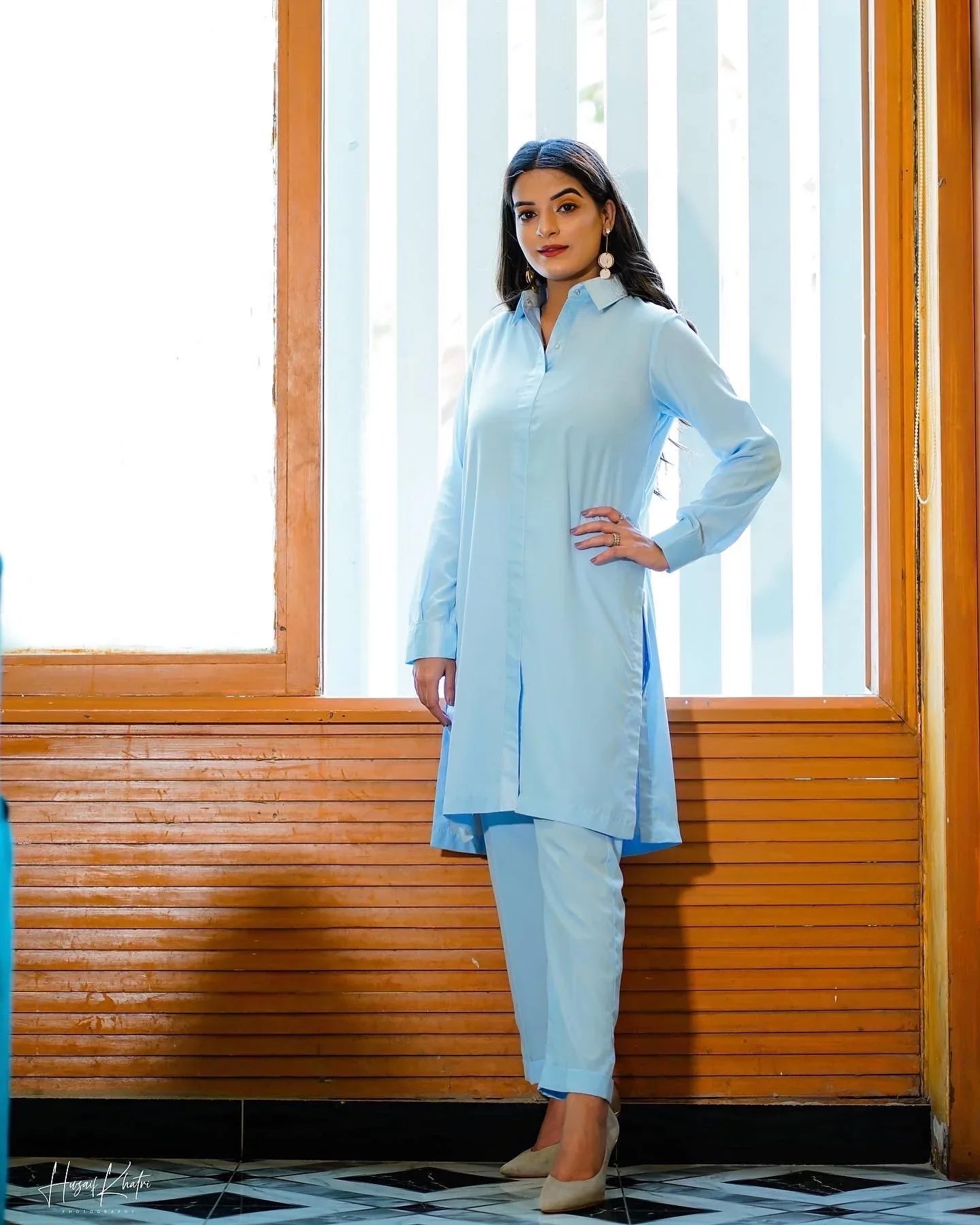 A woman in a sky blue traditional outfit poses confidently indoors. She stands near a window 