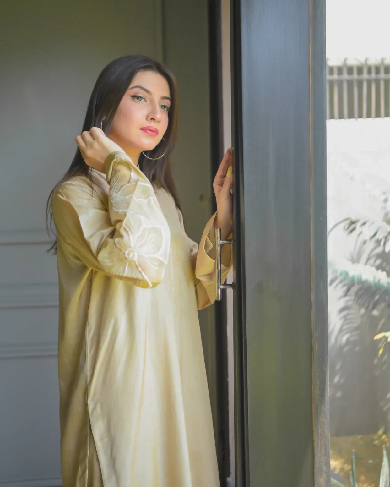 A woman in a beige traditional outfit stands by a large window, gazing outside. Sunlight filters in, illuminating her and nearby plants, creating a serene mood.