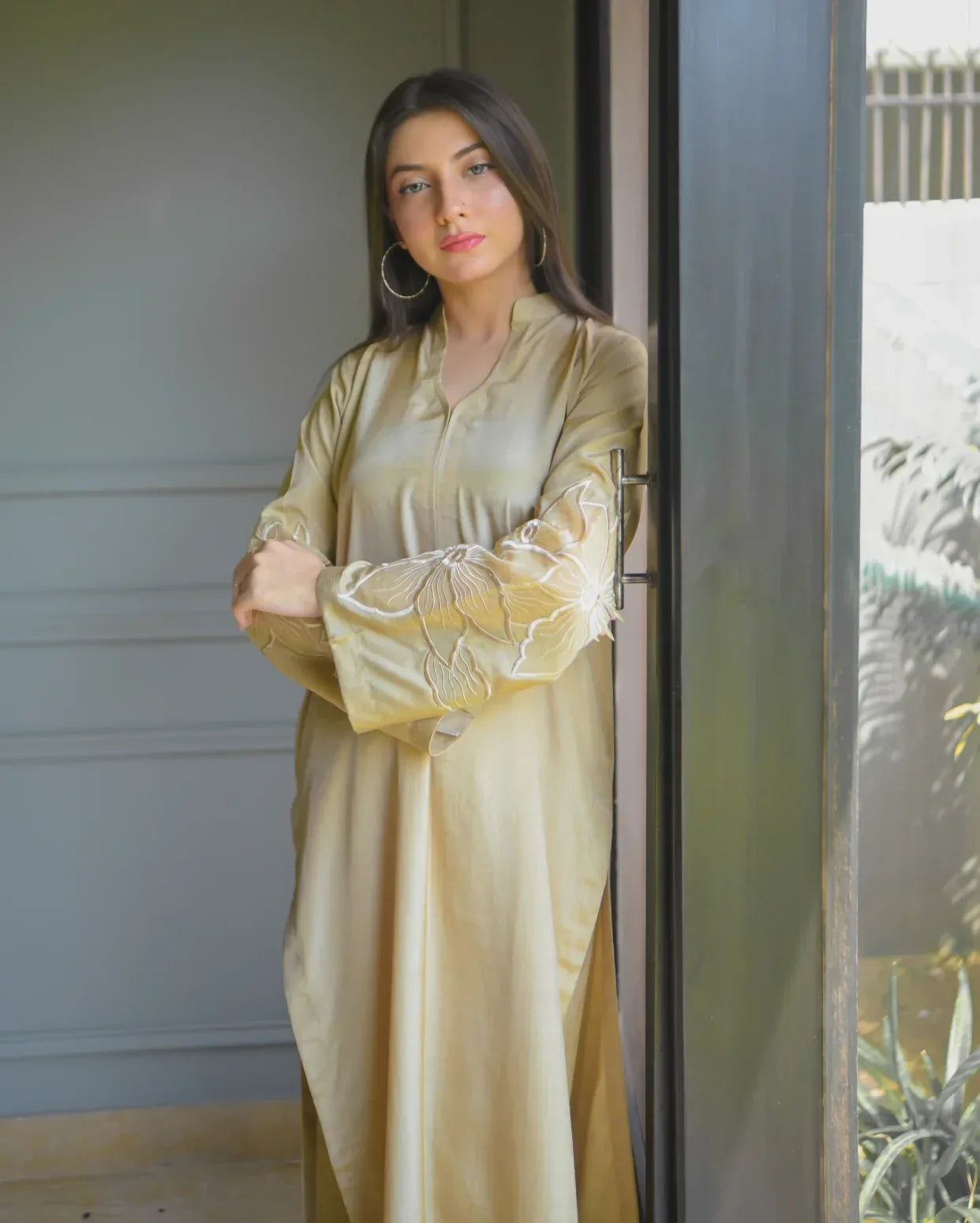 A woman in a beige traditional outfit with embroidery details on sleeves. stands by a large window, gazing outside. Sunlight filters in, illuminating her and nearby plants, creating a serene mood.