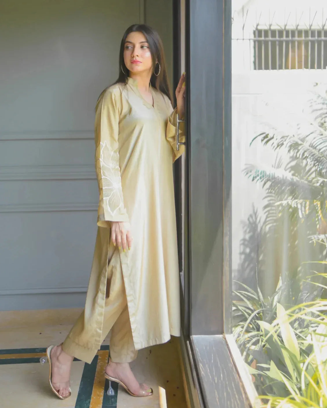 A woman in a beige traditional outfit with embroidery details on sleeves, stands by a large window, gazing outside. Sunlight filters in, illuminating her and nearby plants, creating a serene mood.
