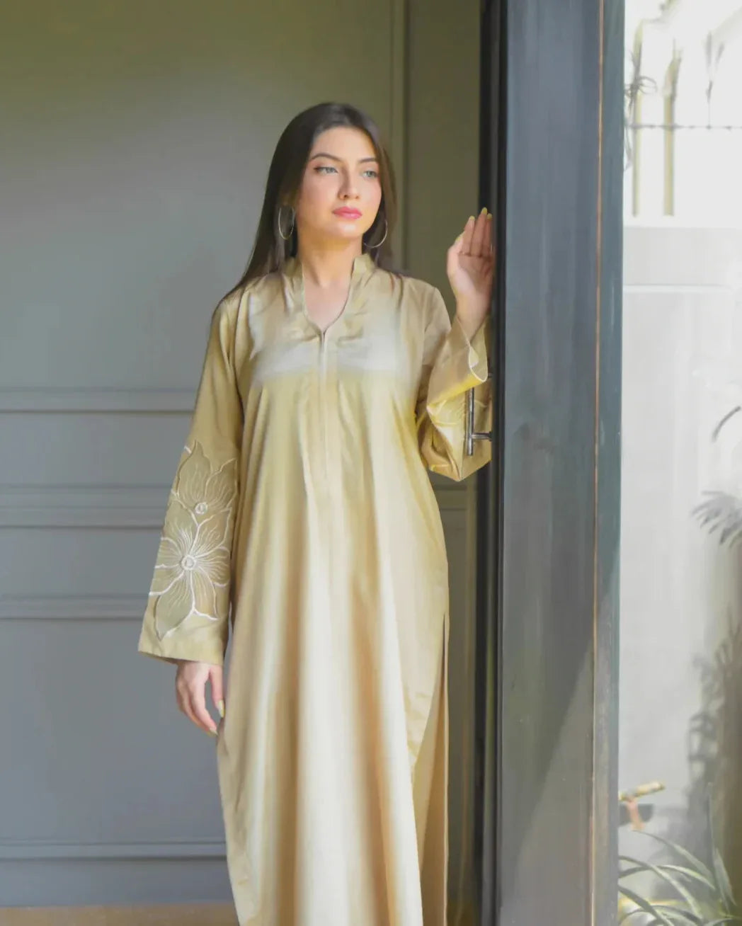 A woman in a beige traditional outfit with embroidery details on sleeves, stands by a large window, gazing outside. Sunlight filters in, illuminating her and nearby plants, creating a serene mood.
