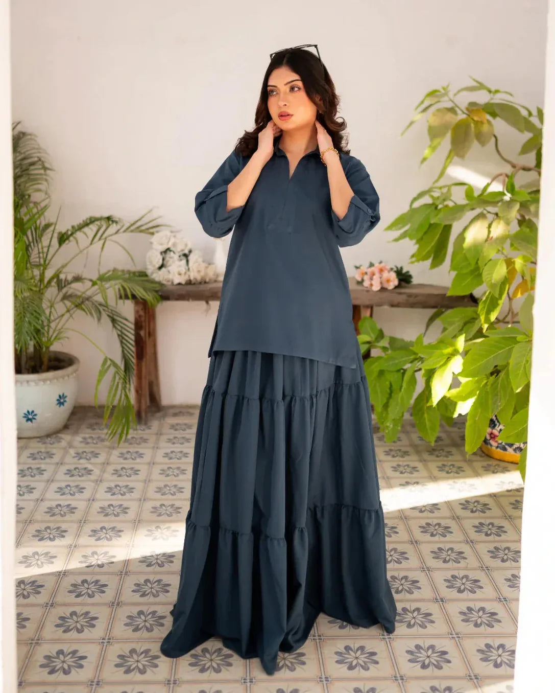 Woman wearing a steel blue skirt and shirt in a room with plants and a tiled floor.
