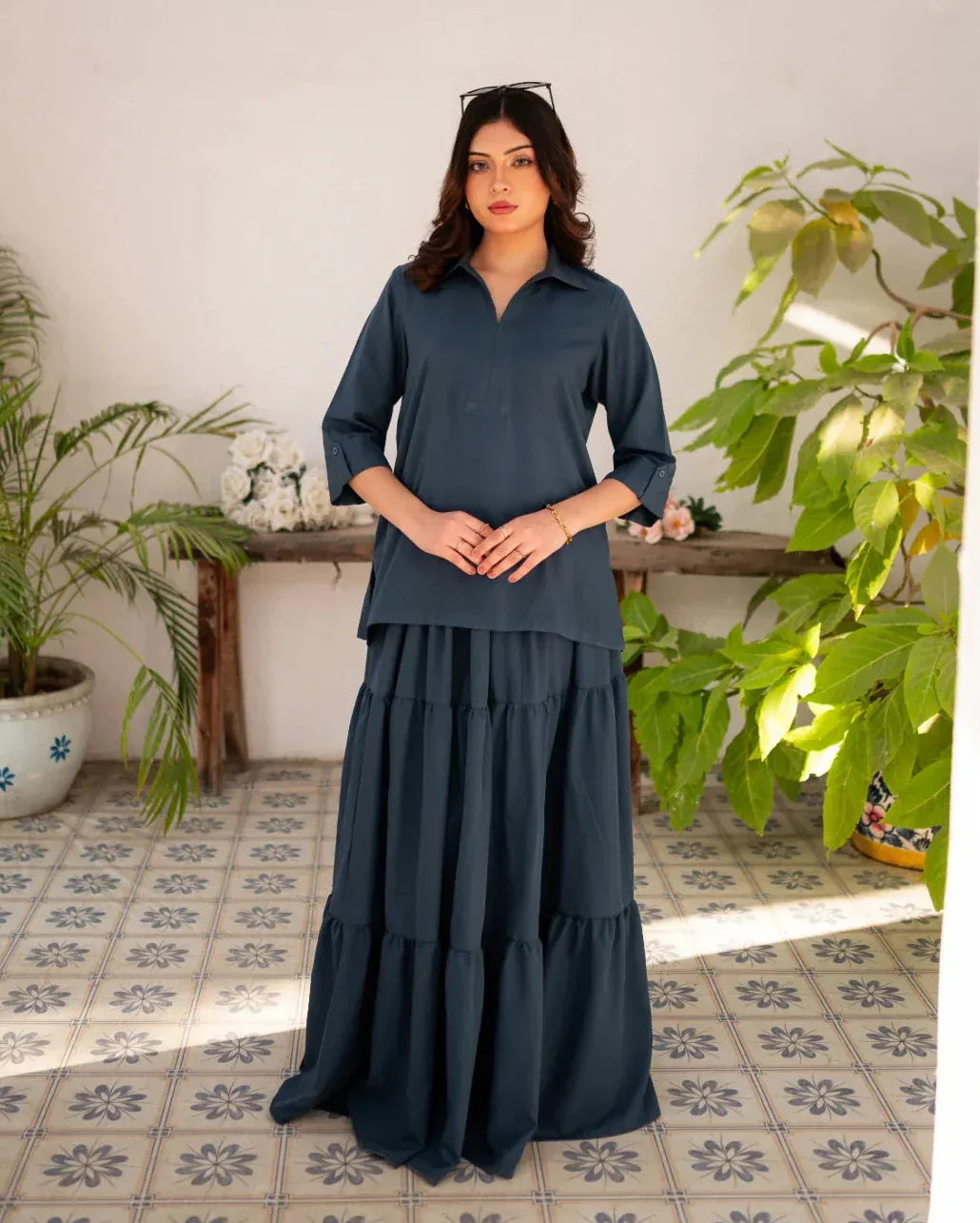 Woman wearing a steel blue skirt and shirt standing in a room with plants and a wooden table.