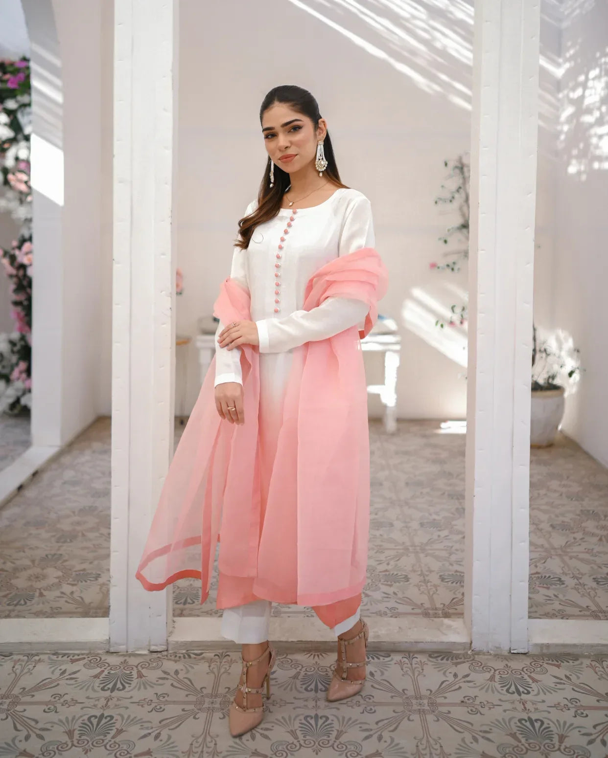 A woman wearing a three piece rawsilk dress with an ombre shirt, trousers, and an organza dupatta in pink and white colors.