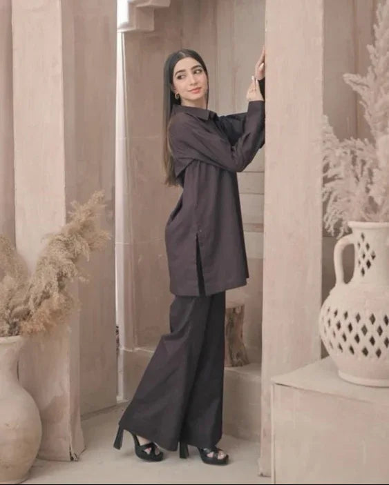 A woman in a stylish dark brown outfit stands in a minimalist room with decorative vases and pampas grass. The ambiance is calm and elegant.