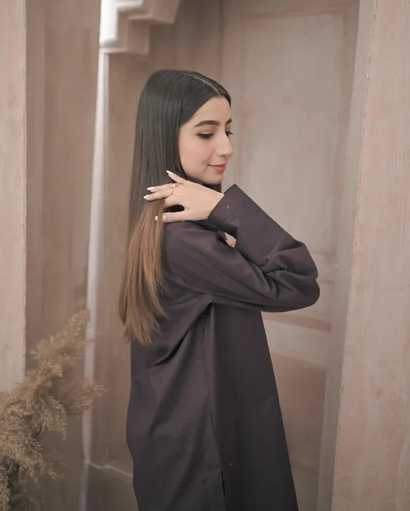 A woman in a stylish dark brown outfit stands in a minimalist room with decorative vases. The ambiance is calm and elegant.