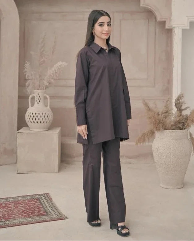 A woman in a stylish dark brown outfit stands in a minimalist room with decorative vases and pampas grass. The ambiance is calm and elegant.
