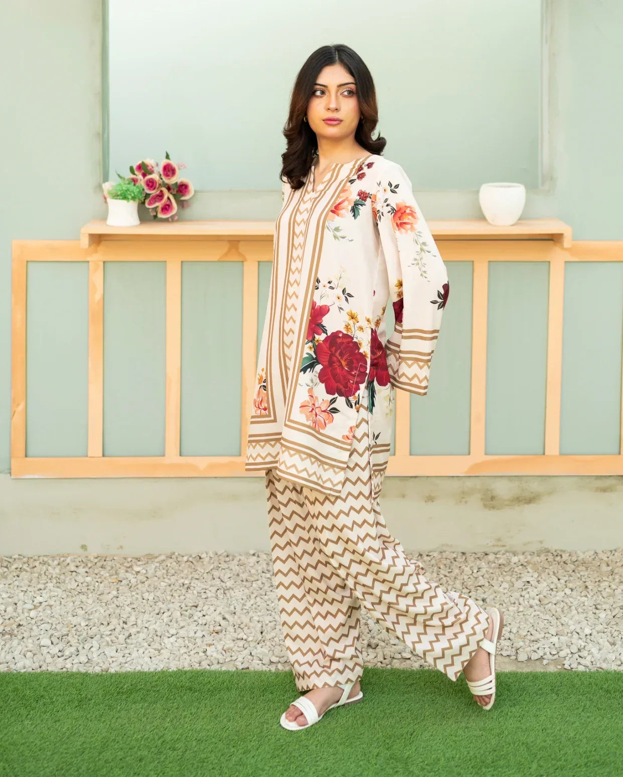 Woman wearing a floral two piece outfit standing on a grassy area with a wooden railing and plants in the background.