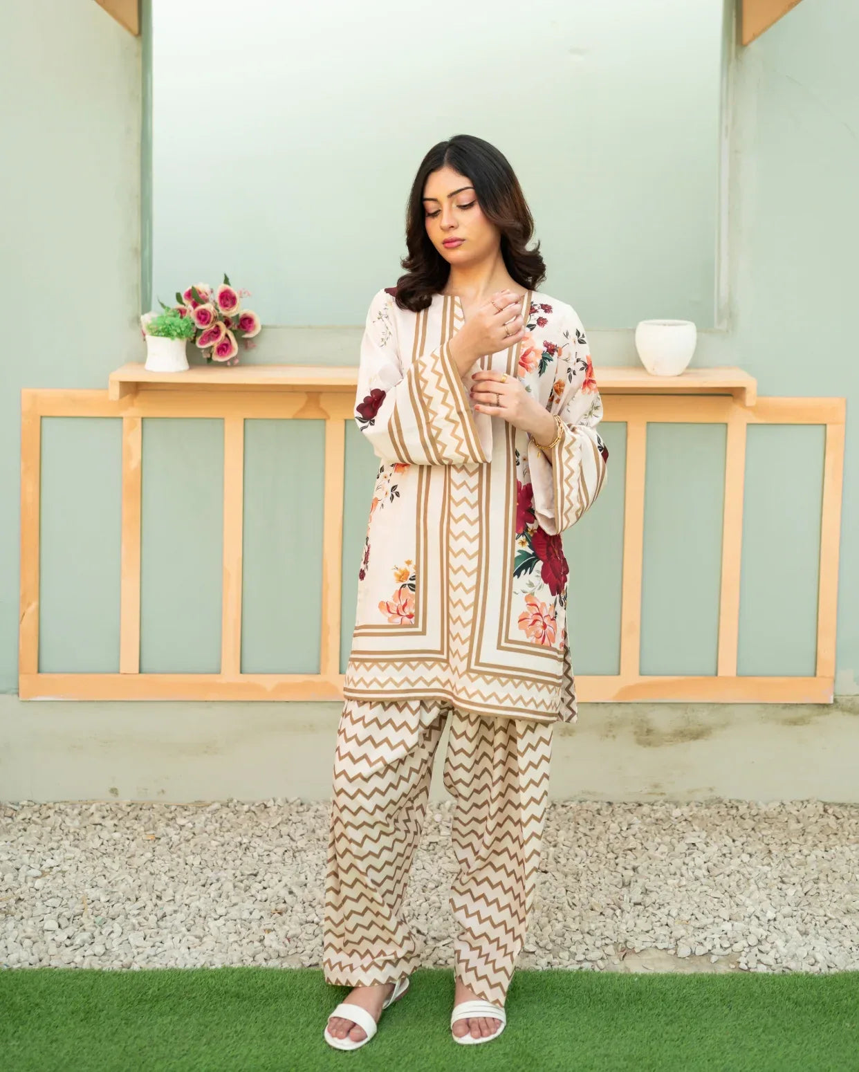Woman wearing a floral printed co ord outfit standing in front of a light green wall with wooden shelves.
