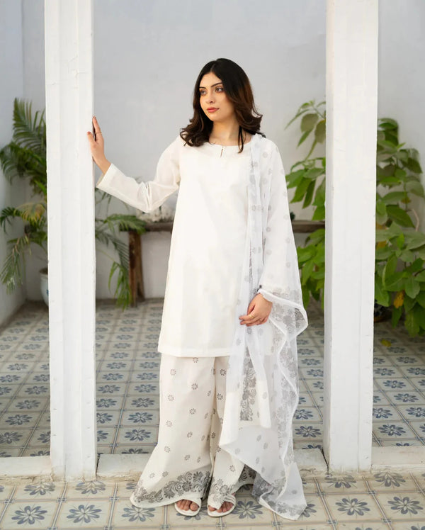 Woman in a white dress, a traditional pakistani kurta set standing in a room with white walls and tiled floor.